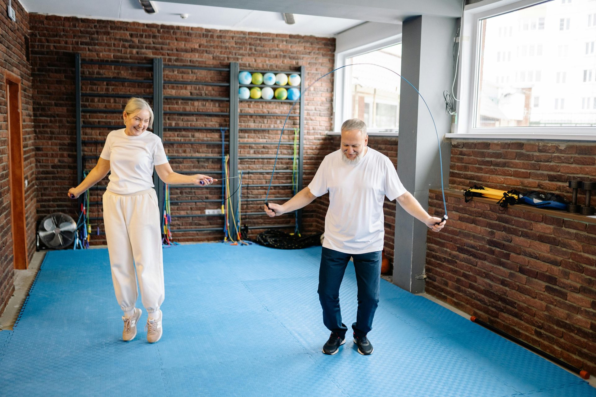 Ein älterer Mann und eine ältere Frau springen voller Freude Seil in einer hellen Turnhalle und werben so für einen gesunden Lebensstil.
