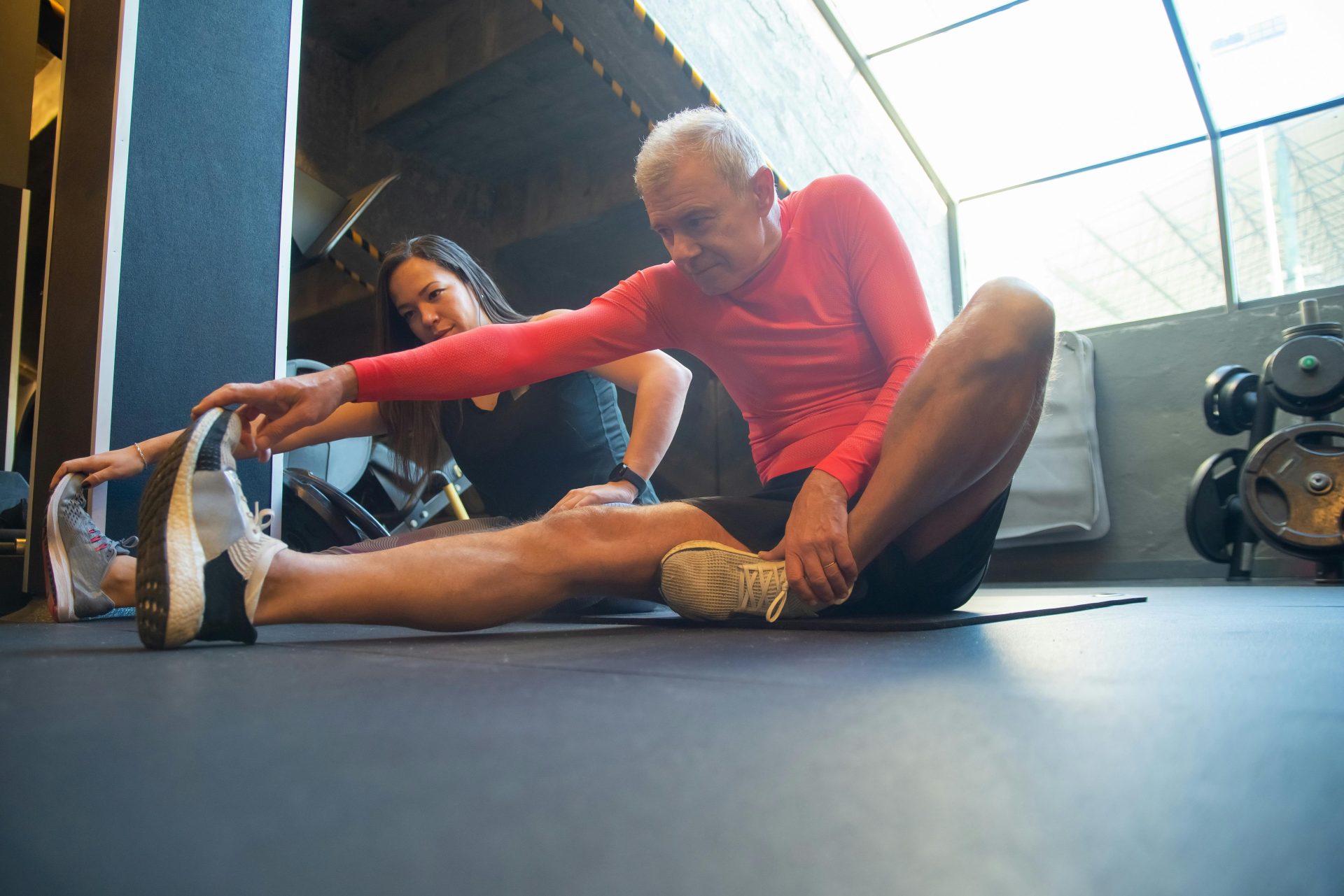 Ein älterer Mann dehnt sich gemeinsam mit einem Trainer in einem modernen Fitnessstudio.