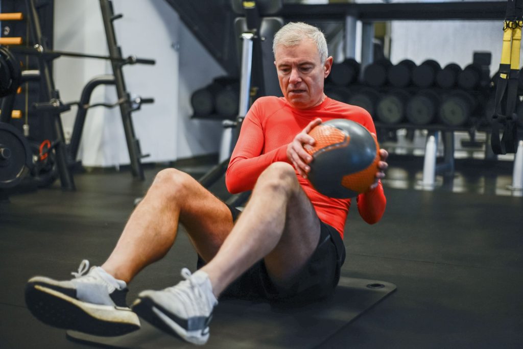 Ein älterer Mann in Sportkleidung führt in einem Fitnessstudio eine Medizinballübung aus. Fördert einen gesunden Lebensstil.