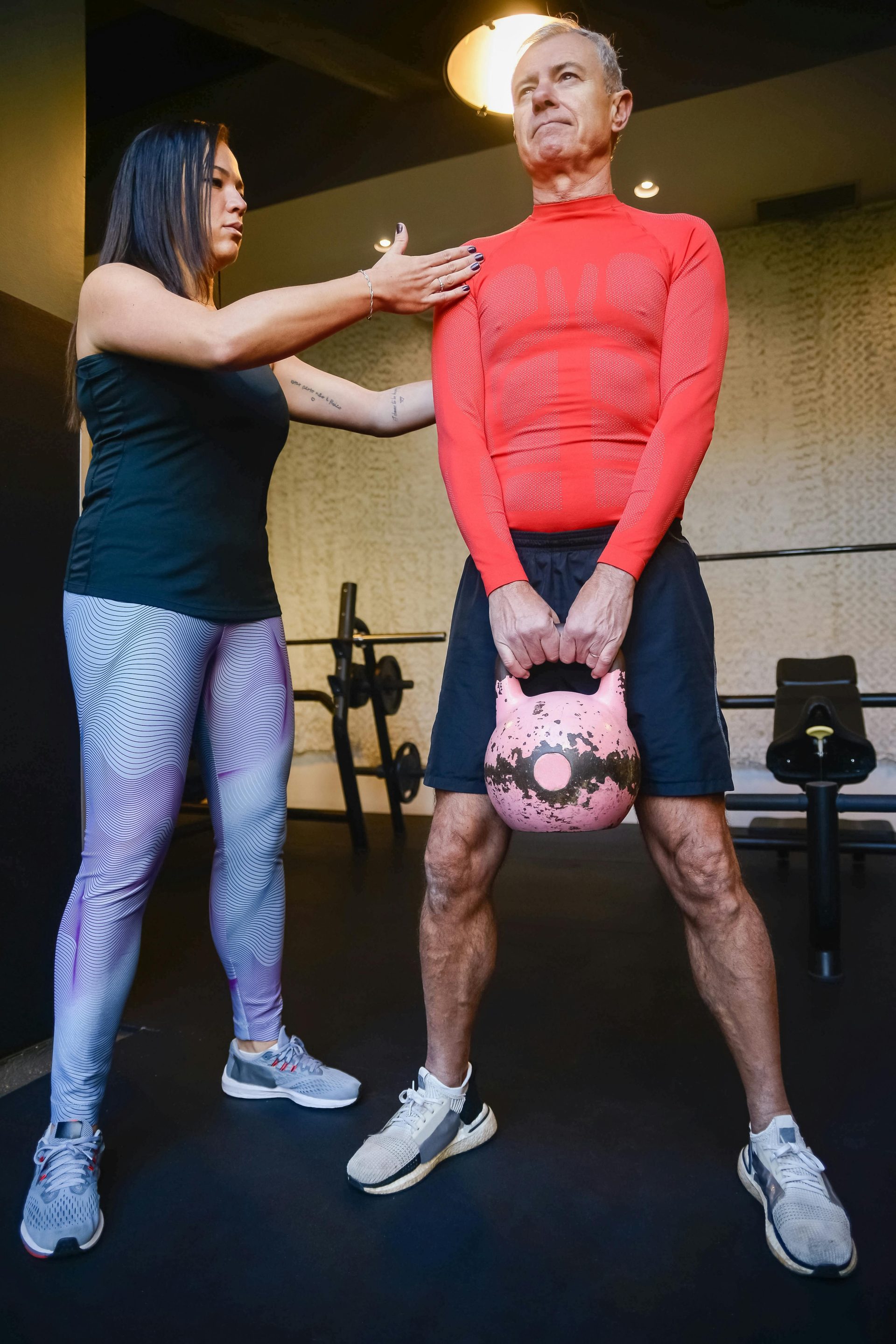 Ein älterer Mann trainiert in einem Fitnessstudio mit einer Kettlebell, angeleitet von einer Trainerin.