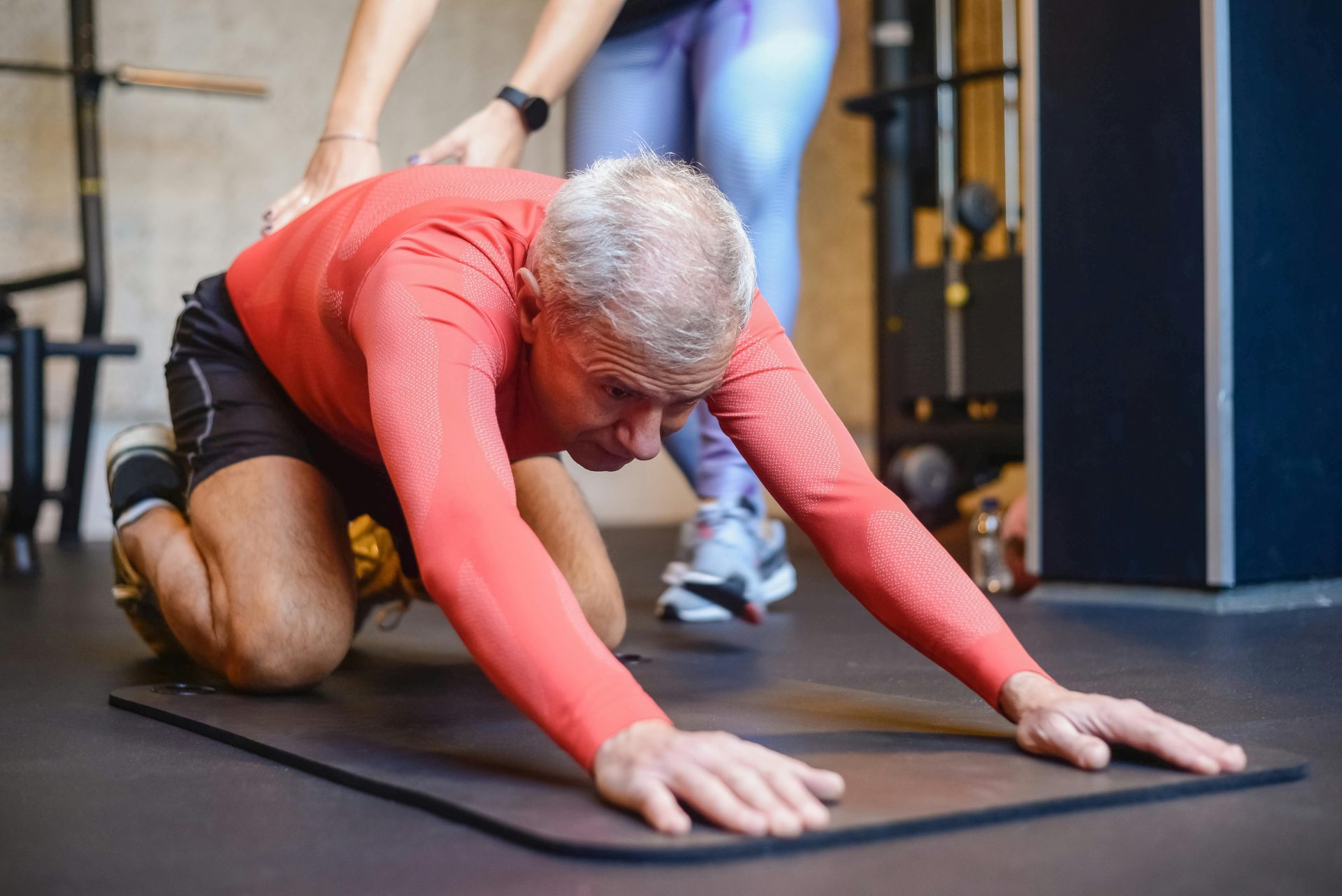 Ein älterer Mann führt in einem Fitnessstudio unterstützte Dehnübungen für einen gesunden Lebensstil durch.